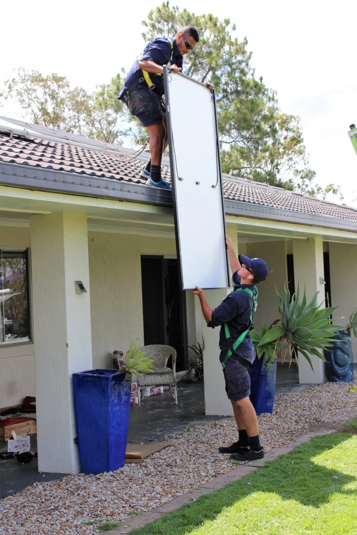 One Ethos Electrical installer on a rooftop passing a solar panel down to a second harnessed installer standing below