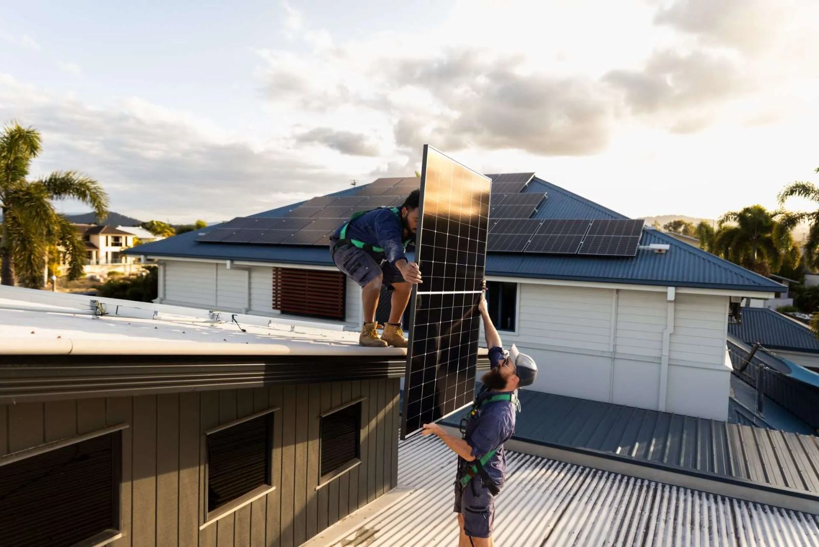 Two harnessed Ethos Electrical installers passing a solar panel between rooftops at golden hour, with a fully-installed panel array visible on the neighbouring roof