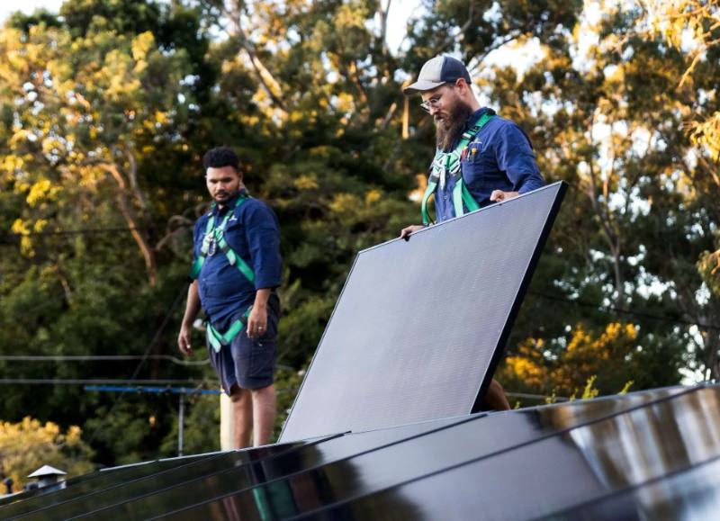 Two installers positioning a solar panel on a roof surrounded by palm trees